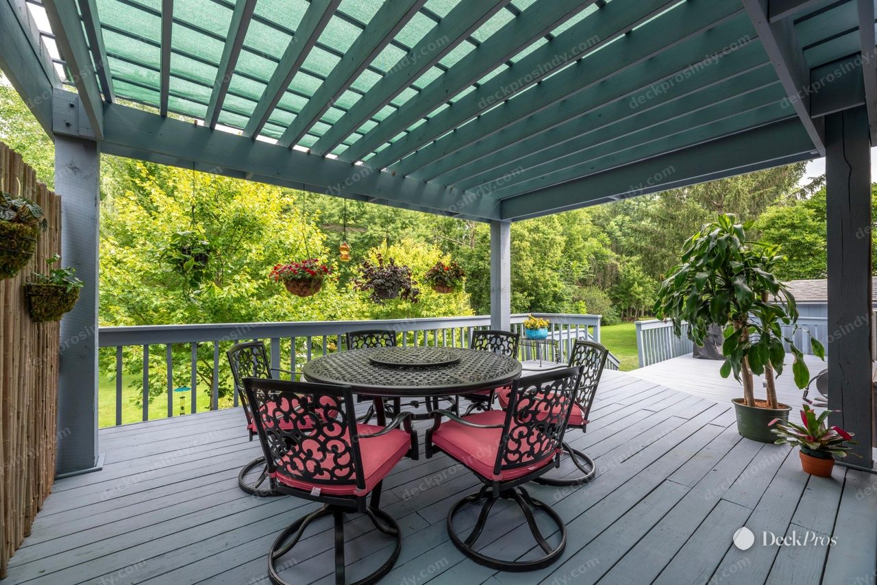 The Spears pergola deck before work: grey-painted decking and blue-grey painted spindle railing under a light blue pergola with shade cloth, with patio furniture and hanging baskets.